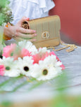 Gold metallic Otis handbag by Elyse & i with gold chain held by a person next to flowers on a blurred background