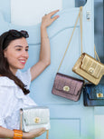 Woman holding a gold Elyse & i Otis handbag with gold chain, surrounded by other Otis handbags on a light blue background