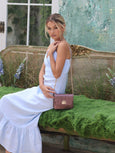 Woman in a white dress sitting on a decorative bench with a green background with a shoulder bag called Otis from brand Elyse & i.