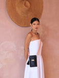 Woman in a white dress holding a black Luna Lock by Elyse & i handbag against a pink wall with a straw hat.