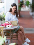 Woman sitting at a table with flowers and an Elyse & i gold Otis handbag, smiling outdoors.