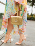 Gold Otis handbag with shell turn lock by Elyse & i held by a person wearing a colorful dress and high heels.