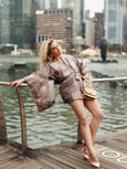 Woman in a patterned outfit leaning against a railing with a Singapore cityscape in the background holding a gold Otis Elyse & i handbag.
