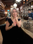 Woman holding a drink in a bar setting with bookshelves in the background and a monochrome snakeprint handbag with shell detail - the Otis by Elyse & i.