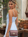 Woman in a light blue dress holding a mulberry metallic handbag by Elyse & i in an outdoor setting with plants and wooden furniture.