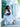 Woman in a white dress holding a blue Elyse & i Nell Knot clutch in an outdoor setting.