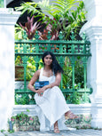Woman in a white outfit sitting in front of a decorative green metal gate with plants holding a navy blue Elyse & i Nell knot bag.
