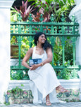 Woman in a white dress standing in front of a green metal fence with plants holding blue Elyse & i Nell knot bag.