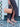 Person holding a tan handbag and wearing beige sandals on a patterned floor.