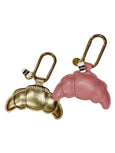 Two keychains shaped like croissants, one gold and one pink, on a white background by Elyse & i