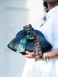 Person holding a black Dawn Scallop leather clutch bag with a patterned green and brown scarf tied around it, wearing a white outfit.