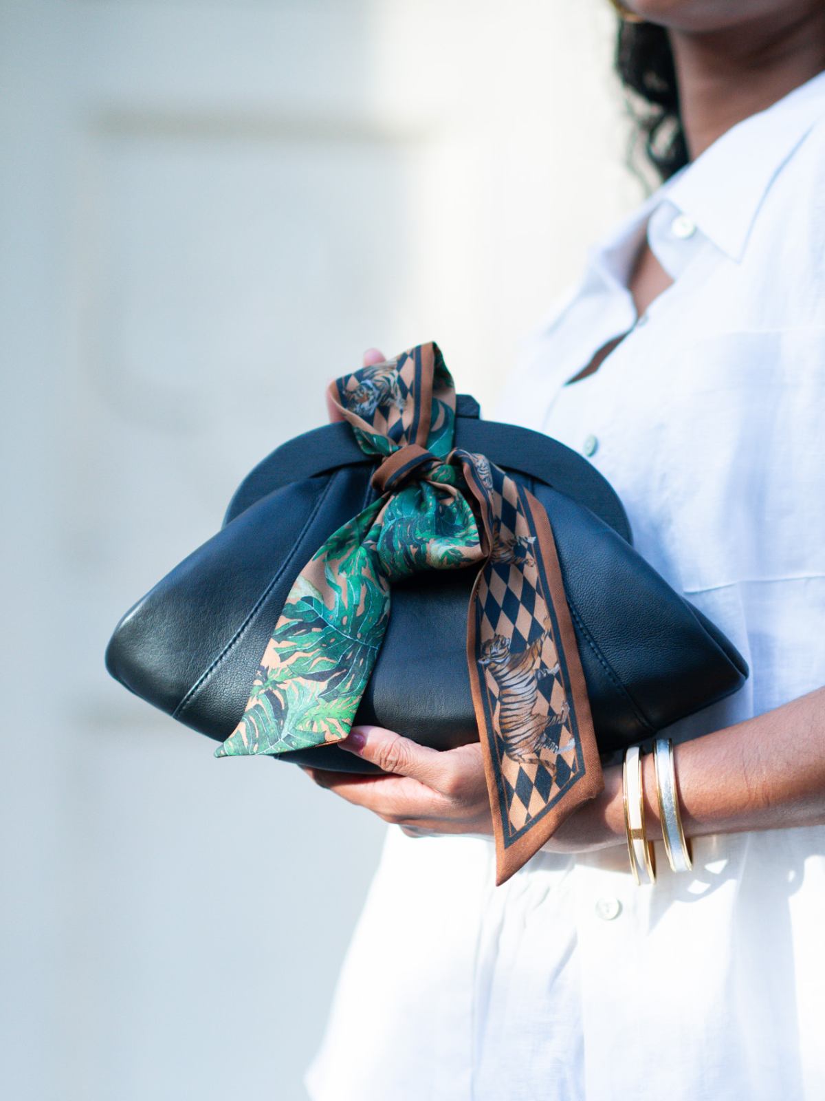 Person holding a black Dawn Scallop leather clutch bag with a patterned green and brown scarf tied around it, wearing a white outfit.