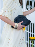 Person holding a black Elyse & i Dawn Scallop clutch with a white lace dress and yellow flower in the background
