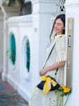 Woman in a white dress with a yellow bow Dawn Scallop black handbag standing next to a white building.