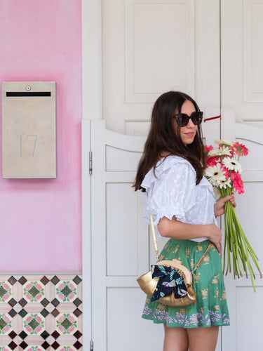 Woman holding flowers and a gold Elyse & i Dawn Scallop clutch in front of a pink wall with a white door.
