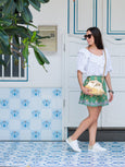 Woman in a white top and green skirt standing in front of a white door with blue accents carrying crossbody the gold Elyse & i Dawn Scallop clutch