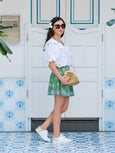 Woman in a white blouse and green skirt standing in front of a white door with decorative blue tiles carrying a gold Elyse & i Dawn Scallop clutch crossbody