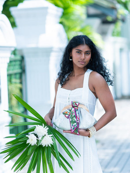 Woman in a white dress holding a floral arrangement with a blurred background, carrying a silver croco embossed leather Dawn Scallop bag with bow by Elyse & i