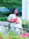Woman in white outfit holding a red Elyse & i Finn Scallop clutch in an outdoor setting with greenery.