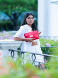 Woman in white outfit holding a red Elyse & i Finn Scallop clutch in an outdoor setting with greenery.