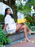 Woman sitting outdoors holding a yellow Finn Elyse & i clutch, surrounded by greenery.