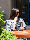 Woman sitting at a table outdoors, holding a black Elyse & i Luna Lock bag with a visible brand logo.