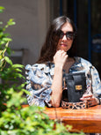 Woman sitting outdoors with a black Luna lock phone handbag, wearing sunglasses and a patterned top.