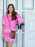 Woman wearing a pink dress with white lace details in front of a white door with blue accents. She's wearing an Elyse & i tawny snakeprint phone bag called the Luna Lock.
