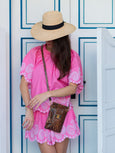 Woman in a pink dress with white lace details and a straw hat, standing against a white wall with blue trim. She's wearing an Elyse & i tawny snakeprint brown phone bag, the 'Luna'.