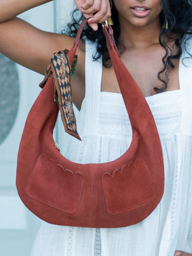 Person holding a brown Elyse & i Martha Suede Hobo bag in terracotta suede with a decorative strap against a light background