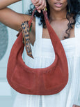 Person holding a brown Elyse & i Martha Suede Hobo bag in terracotta suede with a decorative strap against a light background