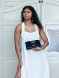 Woman in a white dress holding a black Elyse & i Nell Knot clutch bag in black and gold against a white door background