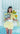 A woman stands in front of a white window and tiled wall, wearing a pale yellow gingham dress with a white waist sash and holding a gray handbag and sunflowers.