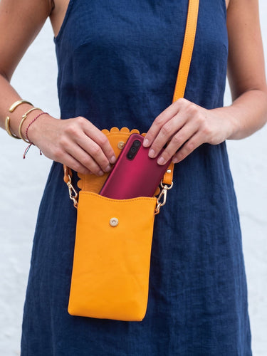 A woman in a navy blue dress holds a bright yellow crossbody phone bag open to show a pink smartphone inside. The bag has a yellow strap, gold-tone hardware, and a scalloped edge.