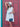 Woman in a white dress and hat holding a blue handbag against a red door with green trim.