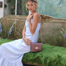 Woman in a white dress sitting on a decorative bench with a green background with a shoulder bag called Otis from brand Elyse & i.
