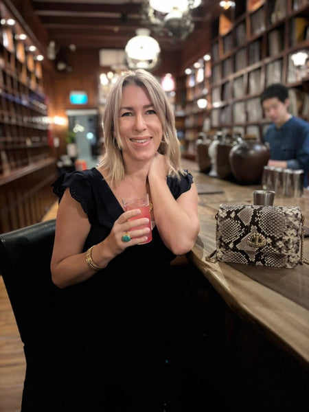 Woman holding a drink in a bar setting with bookshelves in the background and a monochrome snakeprint handbag with shell detail - the Otis by Elyse & i.