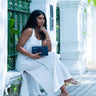 Woman in a white dress holding a blue Elyse & i Nell Knot clutch in an outdoor setting.
