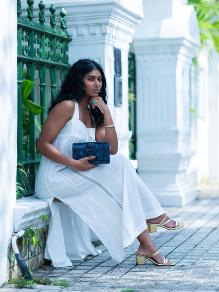 Woman in a white dress holding a blue Elyse & i Nell Knot clutch in an outdoor setting.