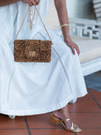 Person holding a cheetah print Finn Scallop clutch bag by Elyse & i with a white dress and gold sandals.