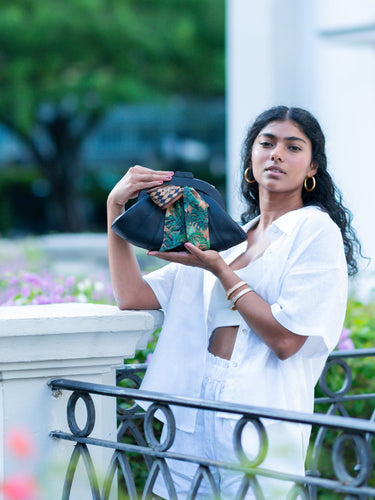 Woman holding a black Elyse & i Dawn bow handbag with a Fleur Kakasi tiger palm silk scarf tied onto it.   