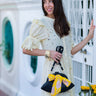 Woman in a white dress with a black Dawn Scallop bag by Elyse & i with a yellow bow standing in front of a decorative white railing.
