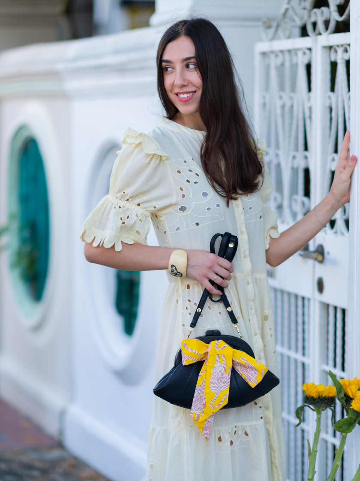 Woman in a white dress with a black Dawn Scallop bag by Elyse & i with a yellow bow standing in front of a decorative white railing.