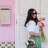 Woman holding flowers and a gold Elyse & i Dawn Scallop clutch in front of a pink wall with a white door.