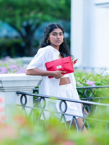 Woman in white outfit holding a red Elyse & i Finn Scallop clutch in an outdoor setting with greenery.