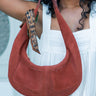 Person holding a brown Elyse & i Martha Suede Hobo bag in terracotta suede  with a decorative strap against a light background