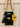 Black handbag with an orange and white strap and a yellow cat-shaped keychain on a light wooden surface.