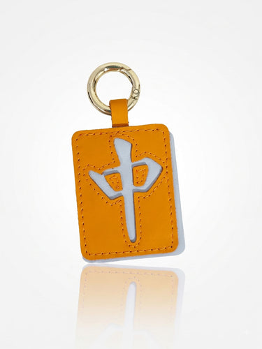 An orange leather luggage tag featuring a white Chinese character 'Zhong' in the center, attached to a gold metal key ring, set against a clean white background with a soft reflection below.