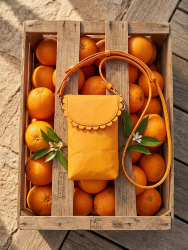 A mustard yellow leather crossbody bag with scalloped trim and a matching strap, styled in a wooden crate filled with oranges on a weathered wood surface.