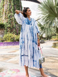 Woman wearing a blue and white tie-dye dress in an outdoor setting with greenery.